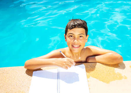 Little boy in swimming pool doing homework holding pen on the border smile look at textbook