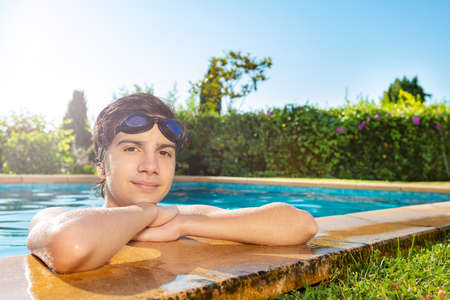 Handsome boy on the border of swimming pool with swim goggles on head look and smileの写真素材
