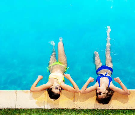 Two happy girls in bikini and sunglasses lay on the border of swimming pool looking at each otherの写真素材