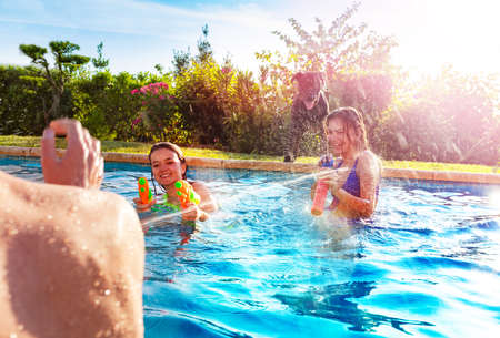 Portrait with two laughing girls in swimming pool shooting water-gun squirt pistol in boys on sunny dayの写真素材