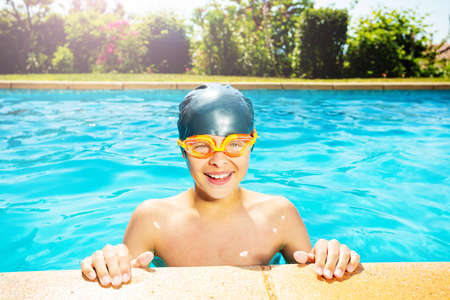 Cute little boy in swimming cap and googles lay on the border of outdoor pool look at camera smilingの写真素材