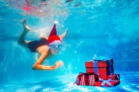 Happy boy in mask dive underwater in Santa Claus hat in the pool reaching for Christmas presents boxes on New Year vacationの写真素材