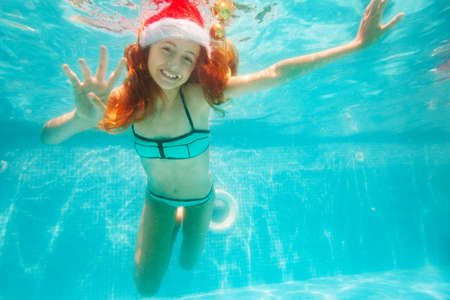 Happy girl dive and swim underwater wearing Santa Claus hat in the pool smile on her New Year vacationの写真素材