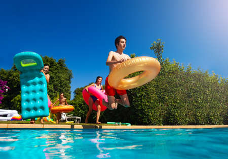 Many of happy teenage kids run to jump into the swimming pool water throw inflatable toys lifting...