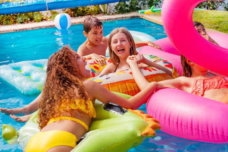 Happy laughing girls swim and float on inflatable toy in a group of friends in swimming pool outside during summerの写真素材