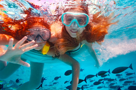 Two beautiful girls in scuba mask swim underwater and wave hand to the camera smiling in the poolの写真素材
