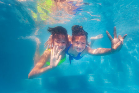 Two happy teenage girls swim together hugging underwater in the pool waving handsの写真素材