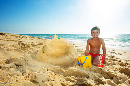 Happy handsome boy build sand castle on the sea beach on vacations sit with shovel and bucketの写真素材