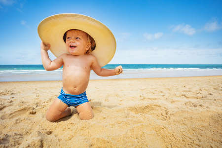 Happy little toddler boy sit on the beach with big straw hat over sea backgroundの写真素材