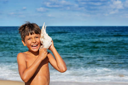 Portrait of a happy relaxed boy listen to huge seashell with big smile over sea backgroundの写真素材