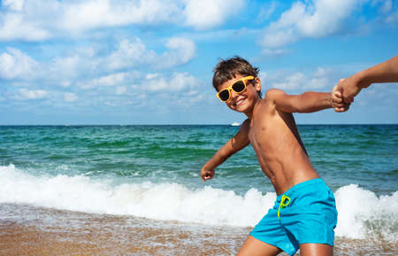 Happy boy in sunglasses pull parents hand to the ocean smiling going to the sea water wavesの写真素材
