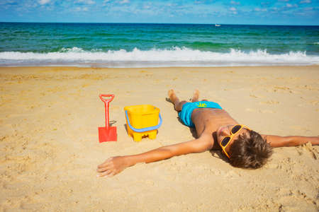 Happy 10 years old boy laying relaxed on the beach with bucket and shovel looking on the seaの写真素材
