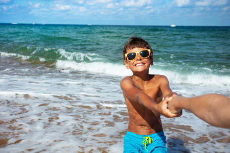 Happy little boy in sunglasses pull parents hand to the sea with big smile and waves on backgroundの写真素材