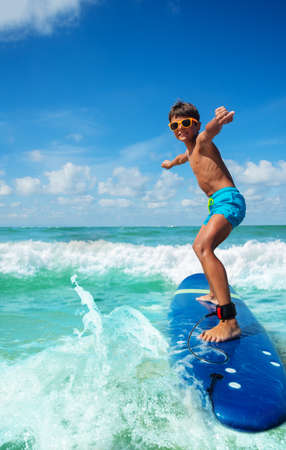 Photo of a boy riding surfboard on the small sea waves practicing with his board in cute sunglasses