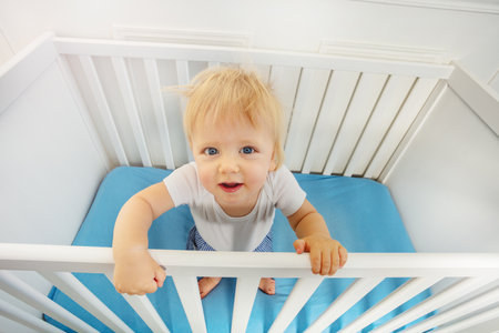 Happy little toddler blond boy stand in baby crib