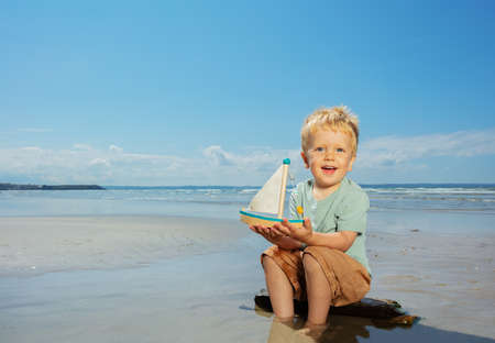 Boy play with toy sail ship sitting on the beachの写真素材