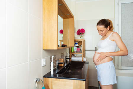 Pregnant woman in bathroom by mirror hold bellyの写真素材