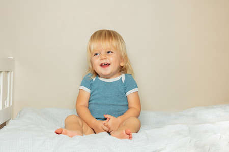 Laughing blond toddler boy sit on the bed in nurseryの写真素材