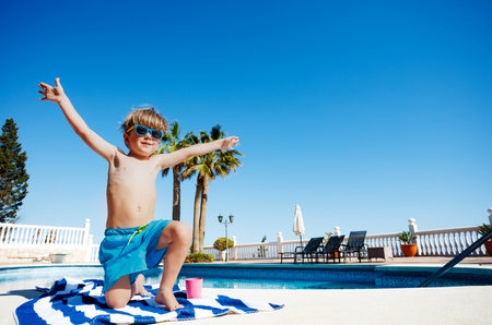 Cute boy in sunshades smiles on towel by pool lifting handsの写真素材
