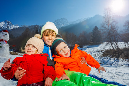 Three kids boys, girl sit together over snowman and mountainsの写真素材