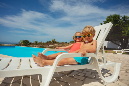Adorable kids in sunglasses chilling by pool on deckchairsの写真素材