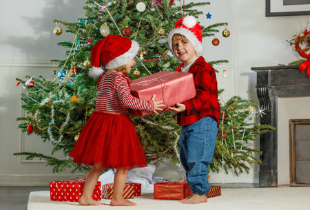 Two kids in festive outfits exchanging gifts by a Christmas treeの写真素材