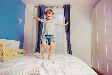 Energetic boy in glasses jumps on the bed at a tidy bedroomの写真素材