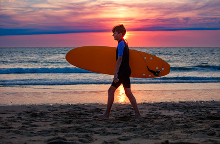 Kid in a wetsuit walk with surfboard by sea at beautiful sunsetの写真素材