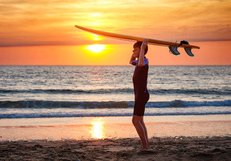 Silhouette of happy surfer kid in wetsuit on the beach at duskの写真素材