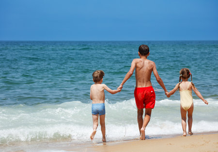 Three kids hold hands, stepping into the ocean waves in summerの写真素材