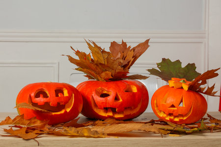 Three carved Halloween pumpkins with autumn leaves on a tableの写真素材