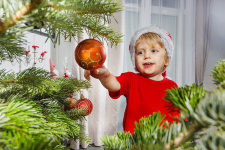 Kid wearing Santa hat places a golden ornament on Christmas treeの写真素材