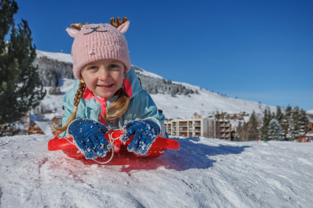 Girl in funny hat sledding on snowy hill under clear blue skyの写真素材