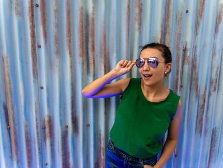 Young beautiful female model poses on a urban stylish metal wall holding her sun glasses and looking to the sky.の写真素材