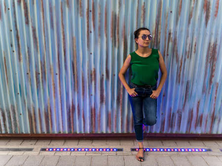 Young beautiful female model poses on a urban stylish metal wall holding her sun glasses and looking to the sky.の写真素材