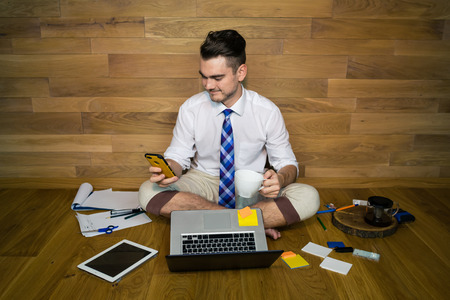 Young man smiling and looking in a smartphone. A barefoot businessman in a funny clothes sits on the floor against a wall with a cup of coffee. Laptop and other gadgets around him.の写真素材