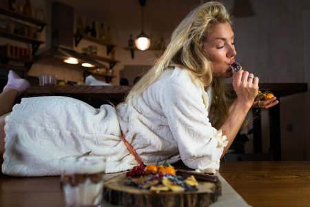 Cute blonde in a bathrobe lying on the table near a beautiful dish on a wooden tray in a stylish apartment in the morning. Young happy woman eats fresh pancakes on the breakfast in the living room at home.の写真素材