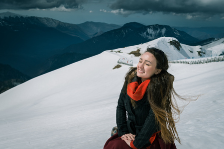 Young woman sits on the top of snowy mountains in the ski resort. Trendy traveler enjoys wonderful day and takes sunbathing. Beautiful mountains and cloudy sky on the backgroundの写真素材