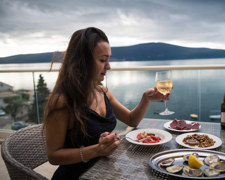 Young pensive woman sits on the balcony at the table with oysters, meat and salad and holds in hand glass with sparkling wine. Elegant girl is ready to eat. Sea and mountains on the backgroundの写真素材