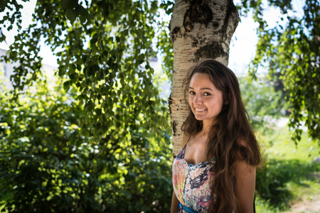 Young attractive woman stands near the tree in the park on the background of green leaves and sun rays.の写真素材