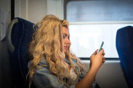 Female passenger in casual clothes sits alone inside the train and ready to departure. Young serious woman looking in smartphone and have fun in trip. Blonde girl travelerの写真素材