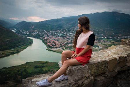 Young stylish woman sits on the stone on background of mountains, river and small city downstairs. Pretty girl looking in side. Travel destination.の写真素材