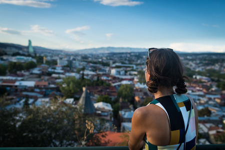 Young unrecognizable woman is standing back at observation deck. View from the hill on old town of Tbilisi. Cityscape in the evening.の写真素材