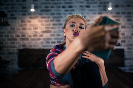 Unusual young woman shows kiss in camera of smartphone at stylish apartment. Portrait of Russian blonde girl taking selfie on the bed in modern bedroom. Stylish interior with soft light.の写真素材