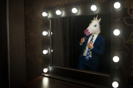 Glamour guy in elegant suit and comical mask looks at himself in the mirror in dressing room.の写真素材