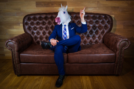 Funny unicorn in elegant suit siting on sofa like a boss and showing hand gesture with raised index finger. Unusual man at home. Freaky guy in mask.の写真素材
