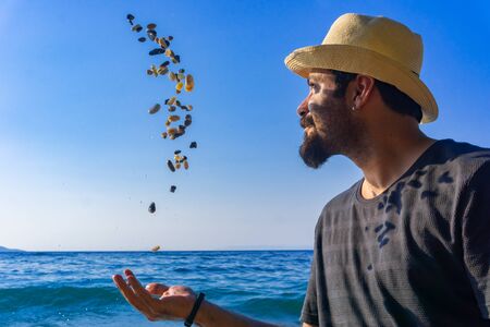 Handsome happy man having fun with pebbles at sunset beach. Man throws beach stones into the air. Summer lifestyle portrait and holiday concept. Young man enjoying and smiling. Izmir, Turkey.の写真素材
