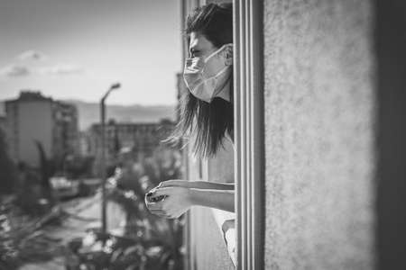 Young woman in medical mask is in self quarantine at home and looking out the window. Covid-19 and home quarantine concept.の写真素材