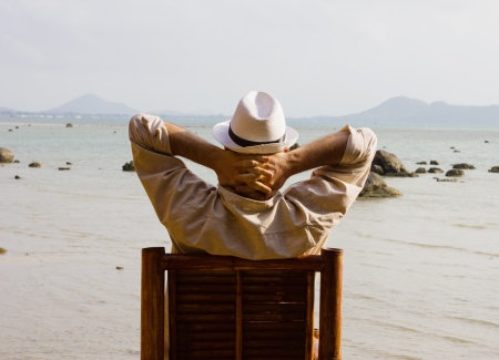 man sitting on a chair and looks at the sea holding his hands on his neck, back viewの写真素材