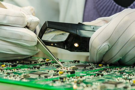 Woman in antistatic gloves holding pincette and magnifier repairing electronic components on PCBの写真素材